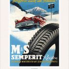 "Bergfest auch im Schnee!" Ein rotes Auto fährt durch Schnee, ein großer Reifen im Vordergrund, Alpenhintergrund.