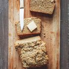 Zwei Scheiben Brot auf einem Holzbrett, eine mit Butter bestrichen; daneben ein Messer.