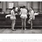 Kalendertag 12. Drei Männer in Ballettkostümen stehen an einer Bar. Im Hintergrund sind viele Flaschen zu sehen.