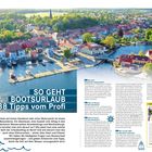 "SO GEHT BOOTSURLAUB - 38 Tipps vom Profi." Ein Hafen mit vielen Gebäuden am Wasser und grüner Umgebung.