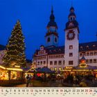 Kalender mit Dezember-Daten. Weihnachtsmarkt bei Abenddämmerung, bunte Lichter, großer Baum und historisches Gebäude.
