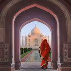 Keine Texte sichtbar. Eine Frau in rotem Sari steht in einem Torbogen und blickt auf das Taj Mahal vor einem Wasserbecken.
