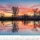 "12 2027" und Wochentage unten. Sonnenuntergang über einem Fluss, Bäume spiegeln sich im Wasser.