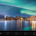 „Norwegen 12“, „Tromsø, Troms, Nord-Norge“. Polarlichter über einer beleuchteten Stadt am Wasser.
