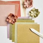 Mehrere bunte Origamipapiere und drei gefaltete Blumen, daneben ein Falzbein.
