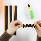 Text: "BASTELKLEBER HOBBY GLUE". Hände falten schwarz-orange Papier, Kleberflasche daneben, Papierstreifen links.