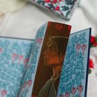 Eine Illustration zeigt das Profil einer Frau mit Kapuze, umgeben von floralen Mustern in Blau und Rot.
