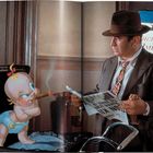 Ein Mann liest eine Zeitung mit der Überschrift „Goofy Cleared“. Neben ihm steht eine Zeichentrick-Babyfigur mit Zigarre.