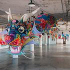 Bunte Dracheninstallation in einem alten Gebäude; leuchtende Farben, detaillierte Muster; industrielle Umgebung aus Beton.