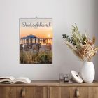 „Deutschland 2027“ steht auf einem Kalender, der einen Steg und Strandkörbe bei Sonnenuntergang zeigt. Darunter eine Kommode mit Deko.