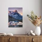 Der Text "Alpen 2027" steht auf einem Wandkalender mit einem majestätischen Bergbild. Darunter ein Tisch mit Büchern und Blumen.