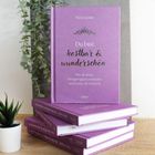 Ein lila Buch mit "Du bist kostbar & wunderschön" von Kira Geiss, steht auf einem Stapel identischer Bücher. Links Pflanze.