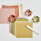 Bunte Papierquadrate, gefaltete Blumen und ein Falzbein auf weißem Hintergrund.