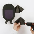 Eine schwarze Papierfledermaus wird mit einer Heißklebepistole zusammengefügt; links ist ein ovaler Körper mit lila Detail.