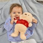 Ein Baby mit blauen Augen hält ein beigefarbenes Kuscheltier mit rotem Oberteil, auf einer grauen Strickdecke liegend.