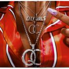 „CITY GIRLS“ auf einer Kette. Eine Hand mit langem, verziertem Nagel zeigt darauf. Roter gestreifter Stoff im Hintergrund.