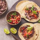 Drei Tacos mit Bohnen, Tomaten und Koriander, dazu eine Schüssel Salsa und Limettenspalten auf einem dunklen Teller.