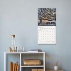 JANUAR 2027, Kalender mit winterlicher Baumlandschaft über einem Tisch auf weißen Regalen mit Karaffe, Büchern und Vase.