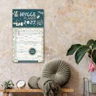 „Hygge Planer 2027“, ein Kalender hängt an einer Wand mit dekorativen Pflanzen und Kissen.