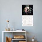 „Januar 2027“ steht auf einem Kalender mit Blumenmotiv, an einer Wand über einem schlichten Regal mit Wasserflasche.