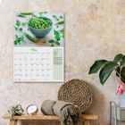 Kalenderblatt mit "Mai" und Bild von grünen Erbsen in einer Schale, an einer strukturierten Wand, daneben Pflanzen und Deko.