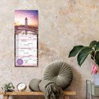 "Familienplaner 2027 Maritime", mit einem Leuchtturm bei Sonnenuntergang als Illustration, hängt an der Wand. Daneben eine Pflanze.
