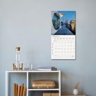 FEBRUAR 2023. Kalender hängt an der Wand über einem Regal mit Wasserflasche, Büchern und Körbchen.