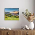 „Zauberhaftes Deutschland 2027“. Ein Kalender an der Wand zeigt eine Landschaft mit Hügeln und einer Stadt im Sonnenuntergang.