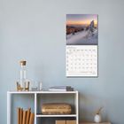 JANUAR 2027 Kalenderseite an der Wand, darüber ein Winterlandschaftsfoto. Darunter ein Regal mit Büchern, Wasserflasche.