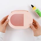 Text: „BASTELKLEBER“
Beschreibung: Zwei Hände halten rosa-ovalförmiges Kartonstück, daneben steht eine Tube Kleber.