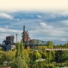 Text: "DIE RHEINSCHIENE – DER WESTEN", "Die Natur erobert Industrieanlagen im Landschaftspark Duisburg-Nord." 

Szene: Industriebauten umgeben von viel Grün.
