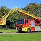 Feuerwehrwagen mit gelber Drehleiter, Umgebung: grüne Wiese, Bäume, blauer Himmel.