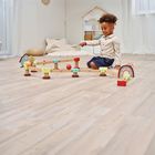 Ein Kind spielt lachend auf einem Holzfußboden mit buntem, kleinem Spielzeughaus und Holzbögen.