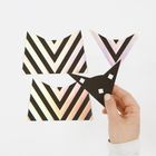 Drei irideszente, schwarz gestreifte Papierstücke. Eine Hand hält ein schwarzes Dreieck mit drei kleinen Quadratlöchern.