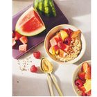 Zahl "65" unten rechts. Zwei Müslischalen mit Früchten; Melonenstücke, Himbeeren, auf einem lila Tablett. Goldene Löffel.