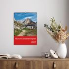 Text: "Hütten unserer Alpen 2027" oben eine Berglandschaft mit Hütte. Rechts Trockenblumen, links Bücher auf Holzregal.