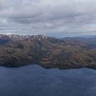 Keine Texte sichtbar. Eine bewaldete Insel in einem weiten, blauen Ozean, umgeben von schneebedeckten Bergen.