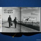 Kapitel II: SHARED STORIES. Zwei Männer auf einem Feldweg. Plakat mit "NEXT TIME TRY THE TRAIN RELAX". Schwarz-Weiß-Foto.
