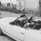 Eine Person mit Sonnenbrille und Mütze sitzt lächelnd in einem offenen Cabrio auf einer ländlichen Straße.