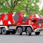 Nummer 6, Rheinmetall, Werksfeuerwehr. Ein großes, sechsrädriges Fahrzeug in rotem Tarnmuster, wahrscheinlich Feuerwehrwagen, steht auf einer Straße vor grünen Bäumen.