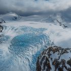 Eine eisige, gebirgige Landschaft mit blauem Gletscher, von verschneiten Hügeln umgeben. Wolken bedecken den Himmel.