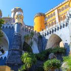 Bunte Burg mit gelben und blauen Türmen, vielen Bögen und Pflanzen im Vordergrund, unter strahlend blauem Himmel.