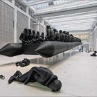 Eine große schwarze Skulptur zeigt ein überfülltes Schlauchboot mit Figuren, in einem modernen, hellen Ausstellungsraum.
