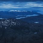 Ein weites Bergpanorama in Blautönen, schneebedeckte Gipfel und dunkle Wolkenformationen.