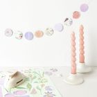 Papiergirlande mit floralen Motiven, zwei gedrehte rosa Kerzen in weißen Haltern, Blumenmuster im Hintergrund.