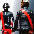 „Daft Punk“-Schriftzug auf Lederjacke, zwei Figuren in Motorradanzügen und Helmen mit roten Gurten.