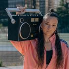 Eine junge Frau in rosa Trainingsjacke trägt einen großen tragbaren Lautsprecher auf der Schulter. Im Hintergrund ein Sportplatz.