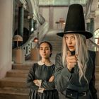 Zwei Frauen stehen vor einer Treppe in einem eleganten Gebäude. Eine trägt einen hohen schwarzen Hut und zeigt nach vorne.
