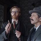 Zwei Männer in älterer Kleidung stehen in einem Raum. Einer hält ein kleines Objekt, der andere schaut konzentriert.