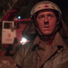 Auf einem Einsatzfahrzeug steht "101". Ein Feuerwehrmann mit rußgeschwärztem Gesicht trägt einen Helm mit rotem Stern.
